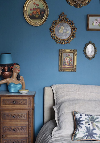 The antique wooden bedside table sits within a modern bedroom with a bright blue wall, kitsch table lamp and a gallery wall of traditional frames. KAte Watson-Smyth looks at ways of introducing brown or rather antique furniture into a scheme. #antiquefurniture #gallerywall #katewatsonsmyth